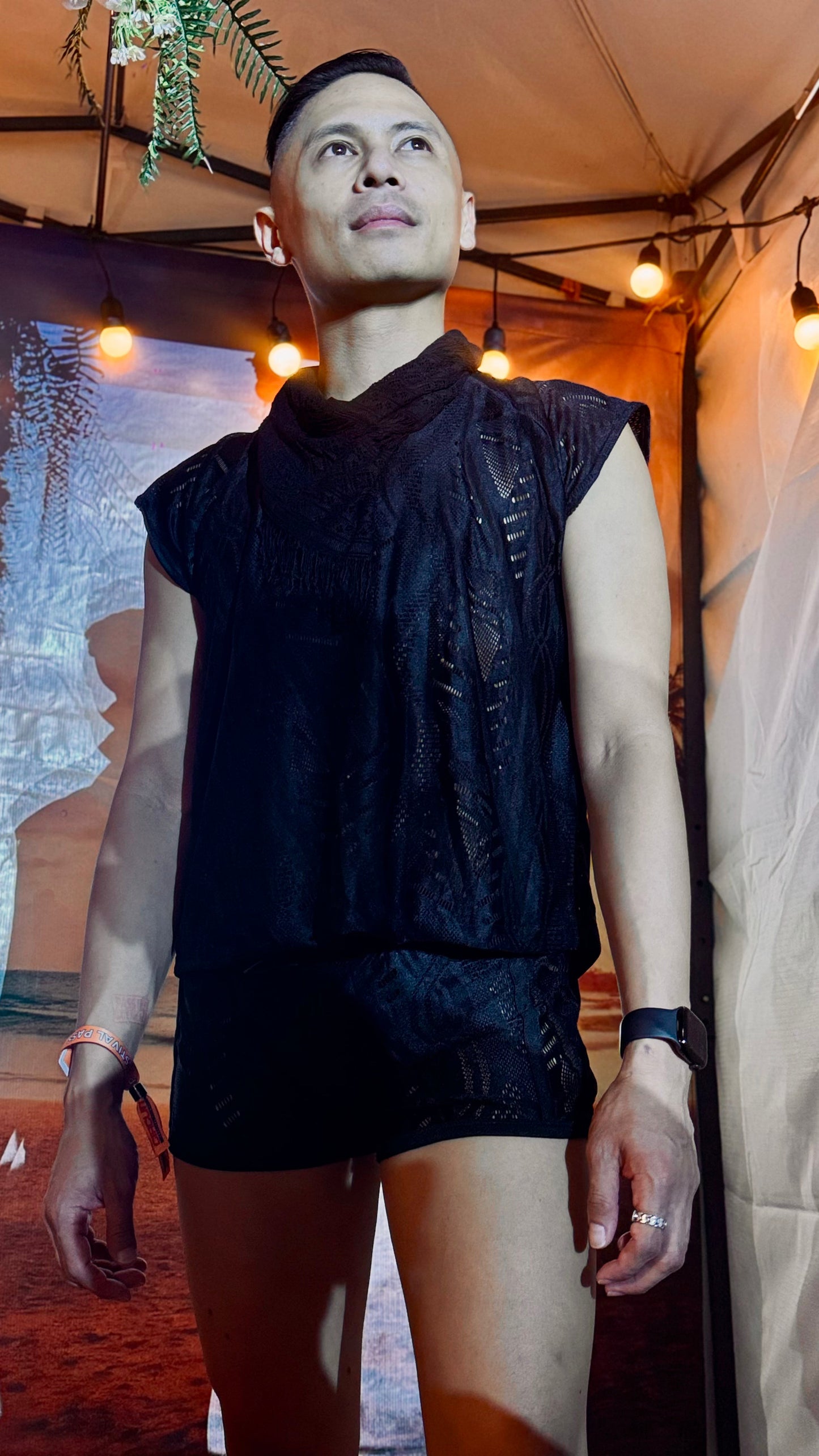 Man wearing the WONGSO Batik Black Lace Festival Sleeveless Shirt with matching shorts under festival string lights