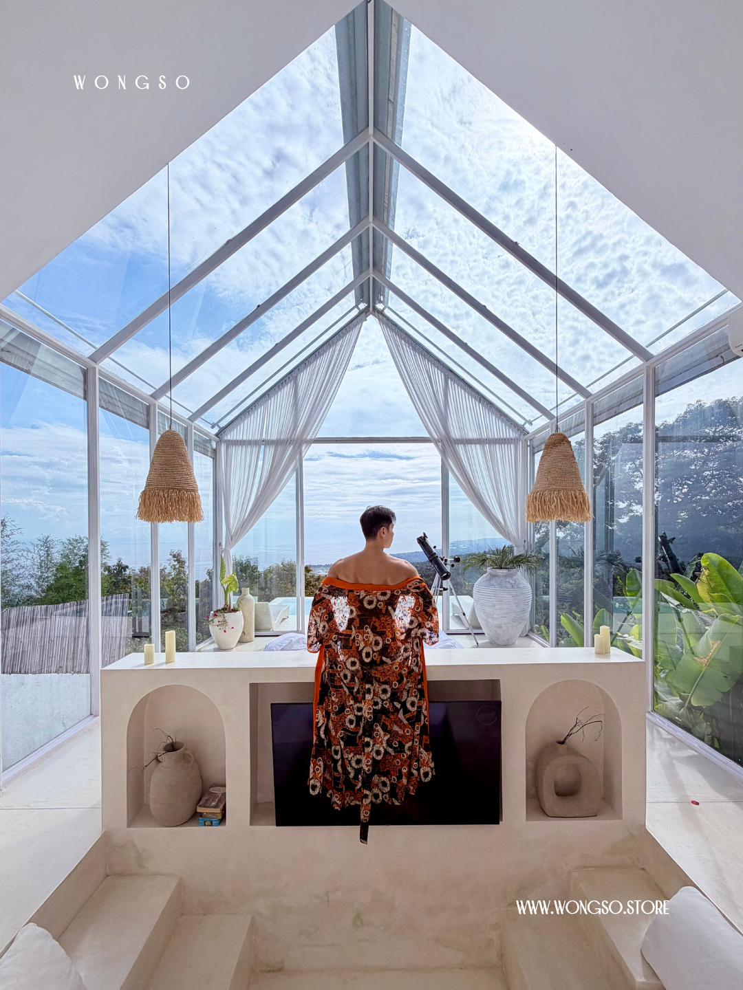 Stefanus, founder of WONGSO, photographed from behind in a floor-length orange floral lace resort kimono worn off the shoulder, looking out to sea from a glass-roofed pavilion in Bali.