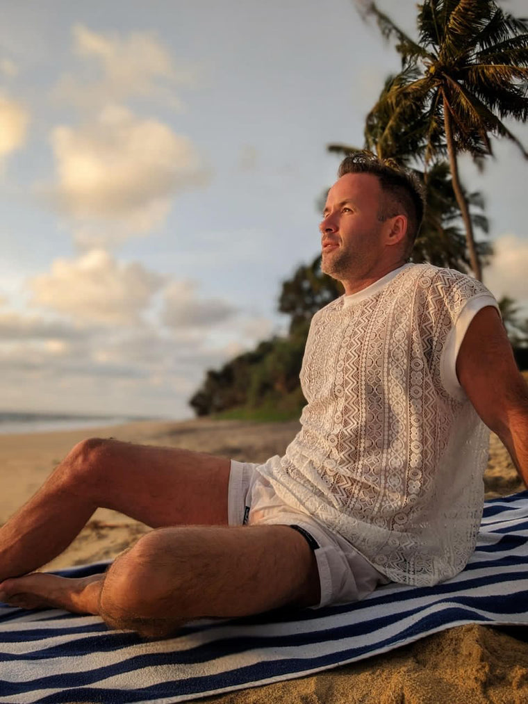 Man in a white lace sleeveless top from WONGSO sitting on a beach towel at sunset under palm trees
