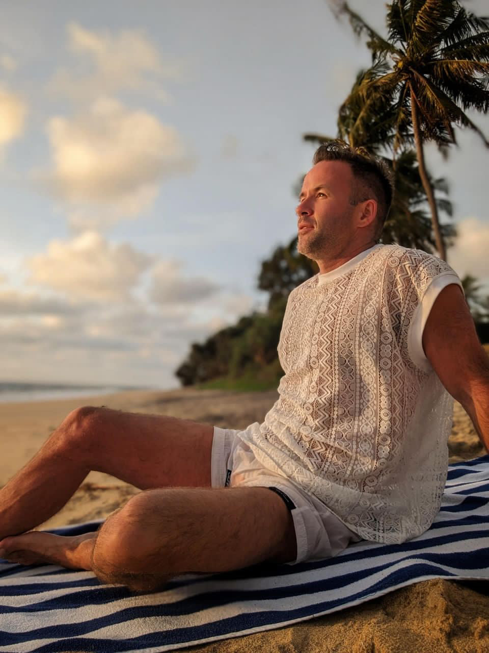 Man wearing the WONGSO White Lace Festival Tank Top on a beach at sunset under palm trees